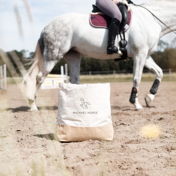 Jutetasche mit schwarzem Motiv auf einem Reitplatz vor einem weißen Pferd